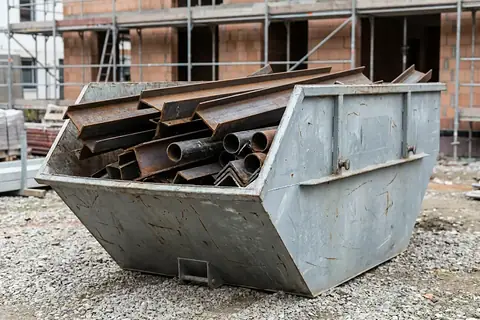 Grauer Absetzcontainer auf einer Baustelle, beladen mit rostigen Stahlträgern, Winkeleisen und Stahlrohren